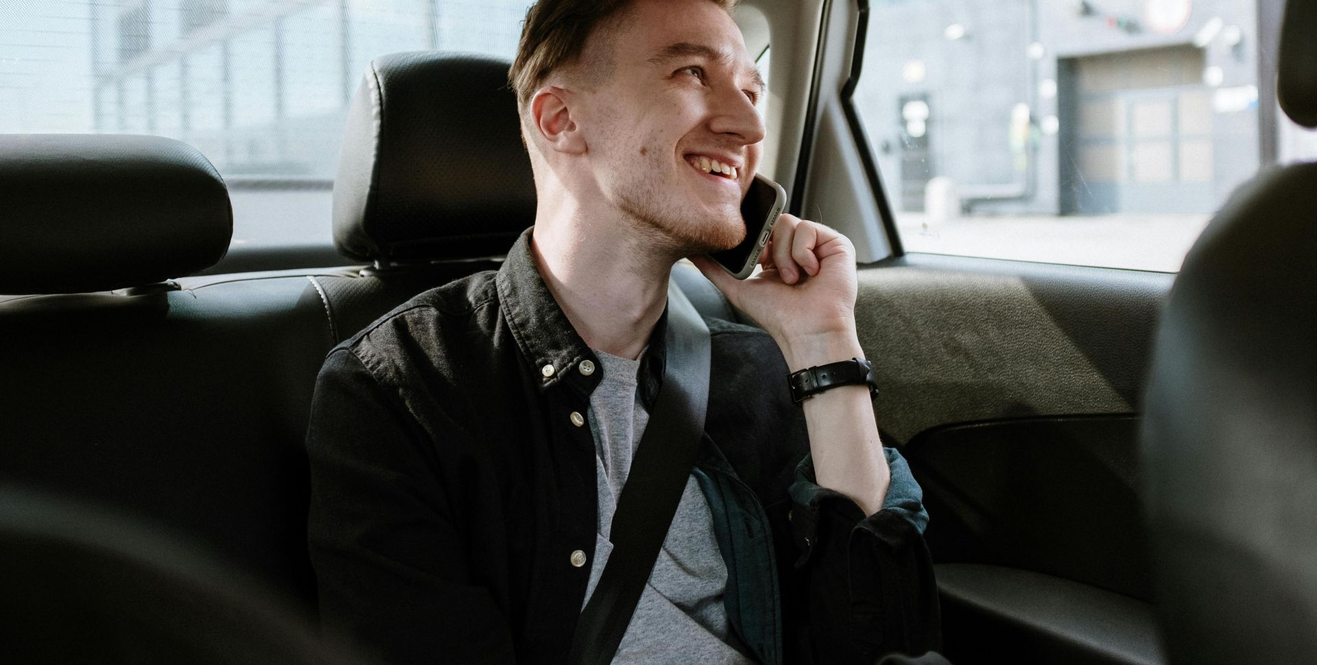 Smiling young man enjoying a phone call while seated in the back seat of a car during a daytime ride.
